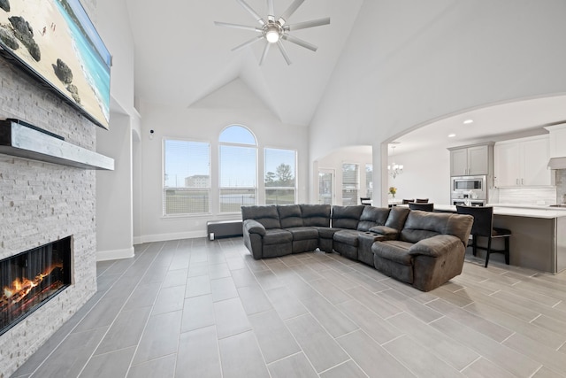 living room with a stone fireplace, ceiling fan with notable chandelier, and high vaulted ceiling