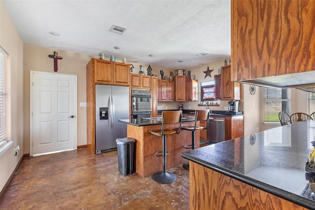 kitchen featuring visible vents, brown cabinetry, dark countertops, appliances with stainless steel finishes, and a breakfast bar area