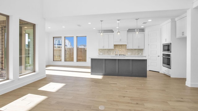 kitchen with oven, a kitchen island with sink, decorative backsplash, white cabinets, and pendant lighting