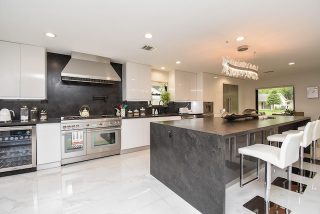 kitchen with range with two ovens, decorative light fixtures, wall chimney range hood, beverage cooler, and white cabinetry