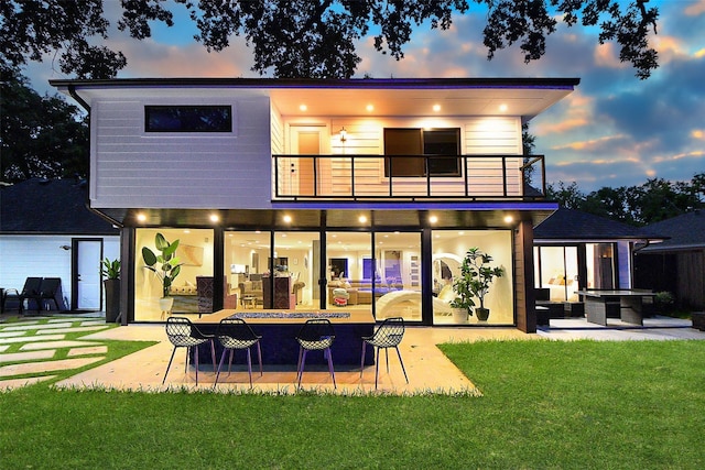 back house at dusk featuring a balcony, a patio area, and a lawn