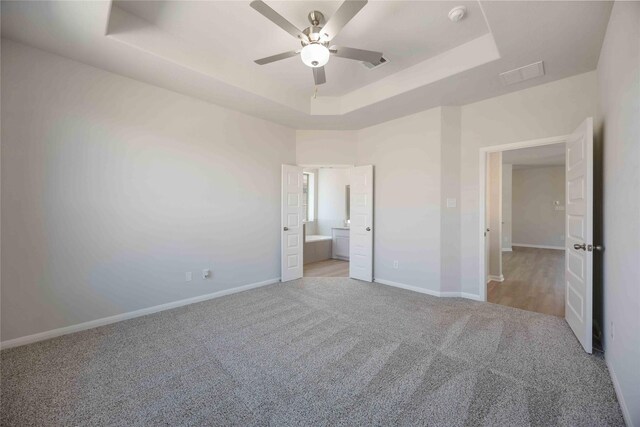 unfurnished bedroom with a raised ceiling, ceiling fan, ensuite bath, and light colored carpet