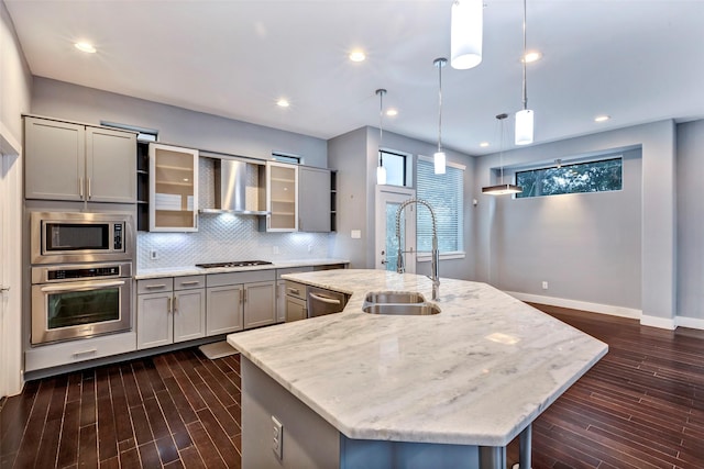 kitchen featuring light stone counters, decorative light fixtures, wall chimney range hood, a kitchen island with sink, and appliances with stainless steel finishes