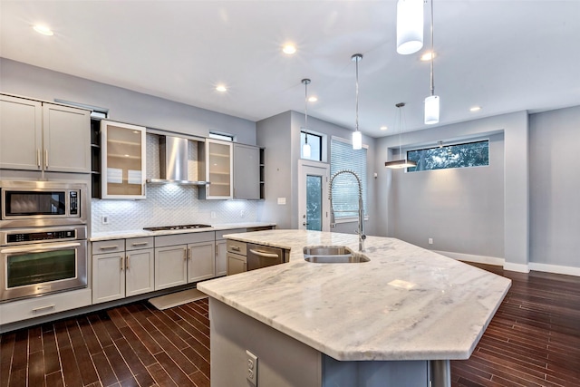 kitchen with stainless steel appliances, a center island with sink, wall chimney exhaust hood, and hanging light fixtures