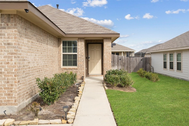 doorway to property featuring roof with shingles, fence, a lawn, and brick siding