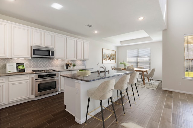 kitchen with stainless steel appliances, a kitchen island with sink, dark stone countertops, white cabinets, and sink