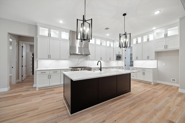 kitchen with wall chimney range hood, pendant lighting, light wood-type flooring, a kitchen island with sink, and white cabinets