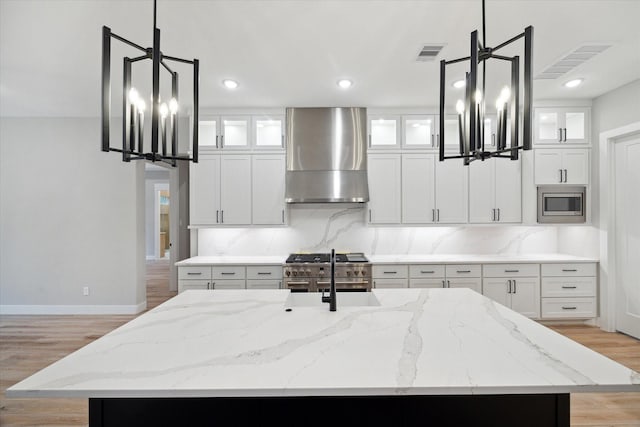 kitchen featuring stainless steel microwave, a kitchen island with sink, wall chimney range hood, and pendant lighting