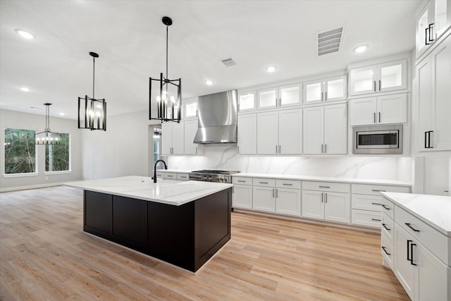 kitchen featuring white cabinets, wall chimney range hood, stainless steel microwave, and a kitchen island with sink