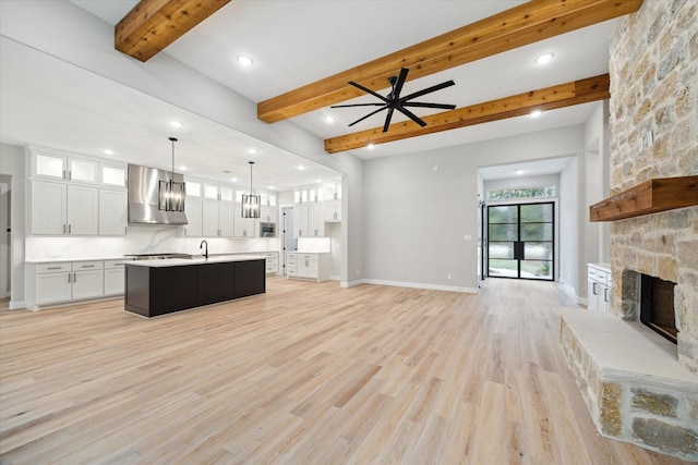 unfurnished living room featuring beamed ceiling, a stone fireplace, sink, light wood-type flooring, and ceiling fan