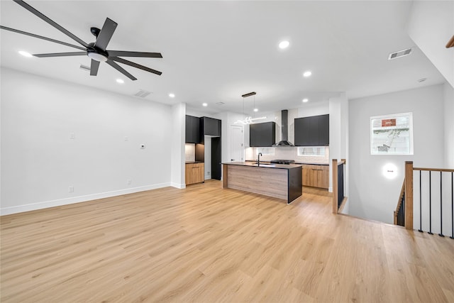 kitchen featuring a center island with sink, wall chimney range hood, tasteful backsplash, light wood-type flooring, and ceiling fan