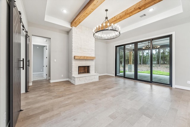 unfurnished living room with a barn door, beamed ceiling, light hardwood / wood-style flooring, a stone fireplace, and a chandelier