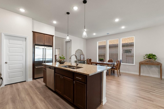 kitchen with sink, stainless steel appliances, light stone counters, hanging light fixtures, and a center island with sink