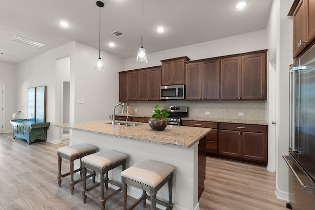 kitchen featuring stainless steel appliances, sink, hanging light fixtures, light stone countertops, and a center island with sink