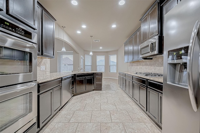 kitchen featuring light stone countertops, hanging light fixtures, decorative backsplash, appliances with stainless steel finishes, and dark brown cabinetry