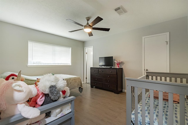 bedroom with ceiling fan, a textured ceiling, hardwood / wood-style flooring, and a nursery area