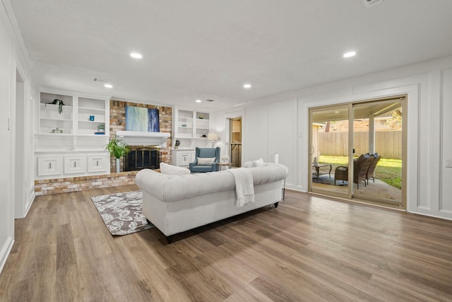 living area with wood finished floors, a brick fireplace, built in shelves, a decorative wall, and recessed lighting