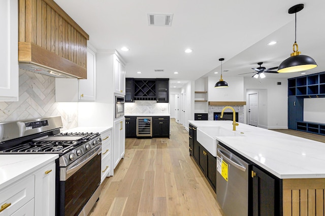 kitchen with wine cooler, decorative light fixtures, appliances with stainless steel finishes, a kitchen island with sink, and white cabinets