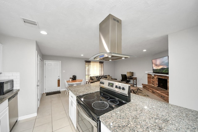 kitchen with appliances with stainless steel finishes, white cabinetry, island exhaust hood, a brick fireplace, and light stone counters