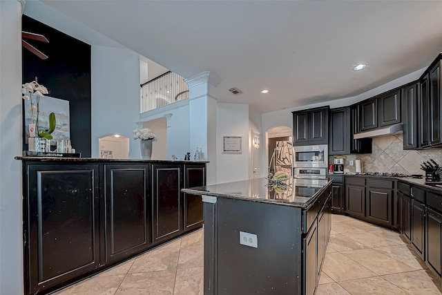 kitchen with light tile patterned floors, appliances with stainless steel finishes, a kitchen island, dark stone counters, and decorative backsplash