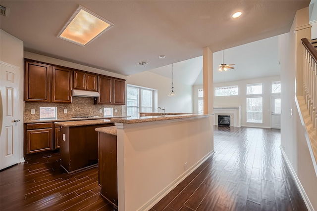 kitchen with dark wood-type flooring, an island with sink, under cabinet range hood, backsplash, and a glass covered fireplace