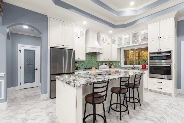 kitchen with stainless steel appliances, a raised ceiling, custom range hood, a kitchen island, and light stone counters