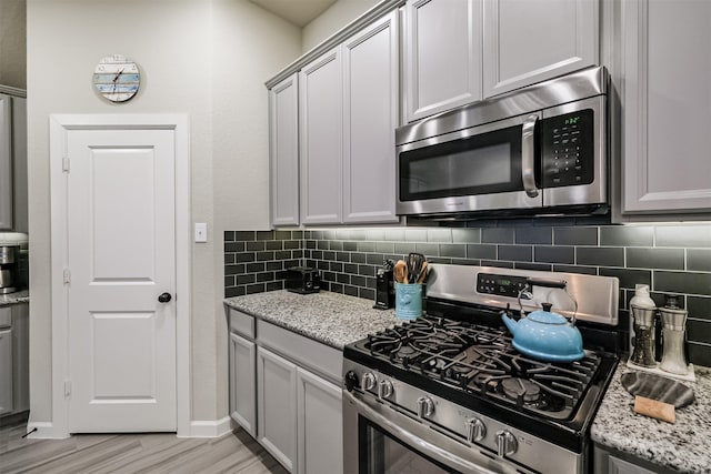 kitchen with gray cabinets, appliances with stainless steel finishes, tasteful backsplash, light wood-type flooring, and light stone countertops