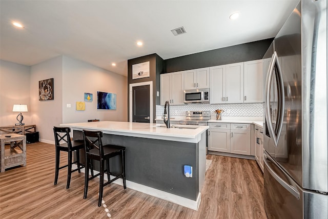 kitchen with light hardwood / wood-style flooring, decorative backsplash, a kitchen island with sink, white cabinets, and appliances with stainless steel finishes