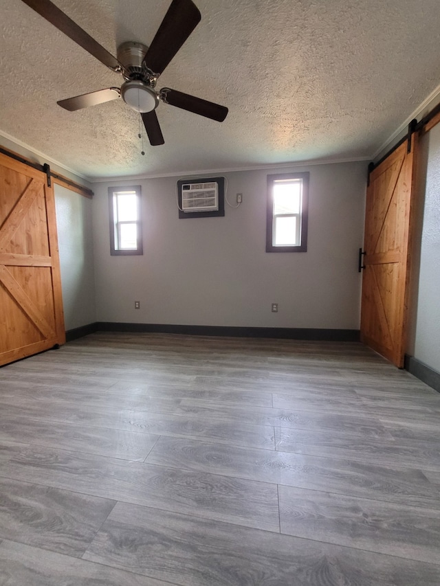 unfurnished bedroom featuring light hardwood / wood-style floors, a barn door, ceiling fan, a wall mounted air conditioner, and a textured ceiling