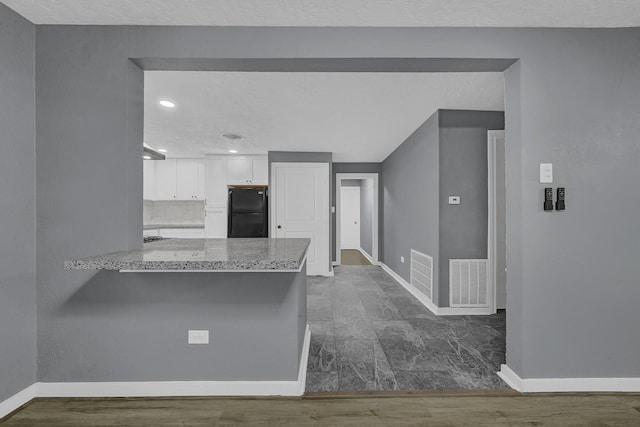 kitchen featuring wall chimney range hood, black fridge, kitchen peninsula, white cabinets, and light stone counters