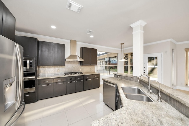 kitchen featuring ornate columns, appliances with stainless steel finishes, wall chimney range hood, pendant lighting, and sink