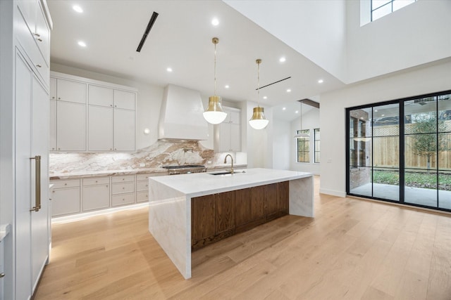 kitchen featuring premium range hood, light hardwood / wood-style flooring, hanging light fixtures, an island with sink, and white cabinets