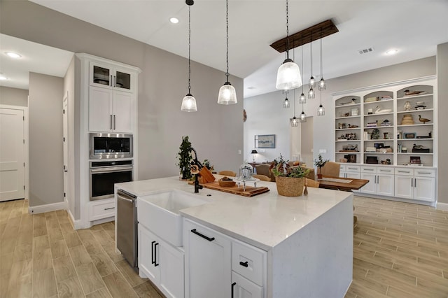 kitchen with an island with sink, appliances with stainless steel finishes, white cabinets, hanging light fixtures, and light stone counters