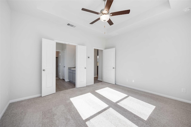 unfurnished bedroom with a raised ceiling, light colored carpet, ceiling fan, and ensuite bath