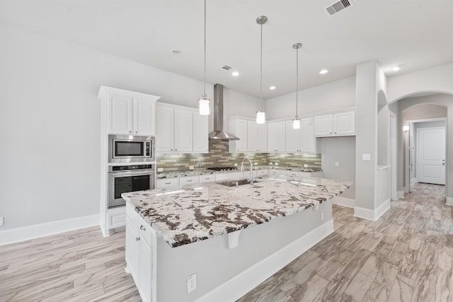 kitchen with appliances with stainless steel finishes, a kitchen island with sink, white cabinetry, and wall chimney range hood