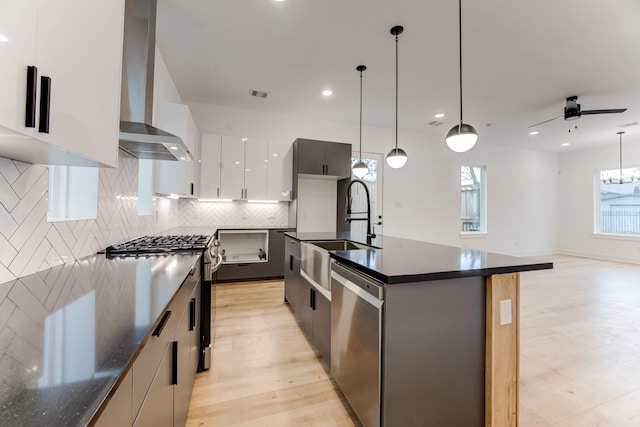 kitchen with wall chimney range hood, pendant lighting, a kitchen island with sink, appliances with stainless steel finishes, and white cabinets