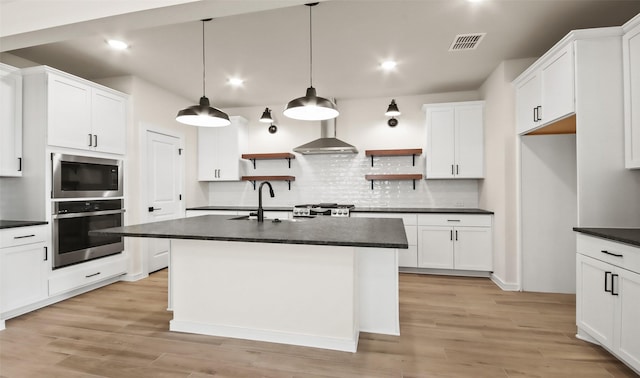 kitchen featuring wall chimney exhaust hood, hanging light fixtures, a kitchen island with sink, oven, and built in microwave