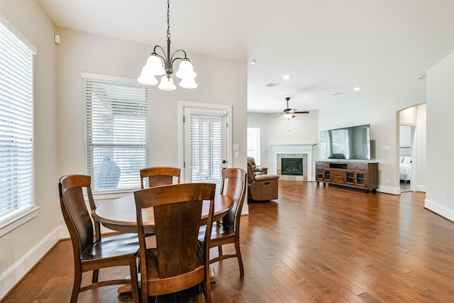 dining room featuring dark hardwood / wood-style flooring, a wealth of natural light, ceiling fan with notable chandelier, and a tiled fireplace