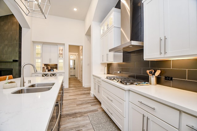 kitchen featuring stainless steel gas cooktop, wall chimney range hood, white cabinets, light stone counters, and sink