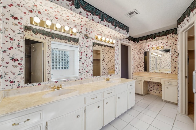 full bathroom featuring a walk in closet, double vanity, a sink, a textured ceiling, and wallpapered walls