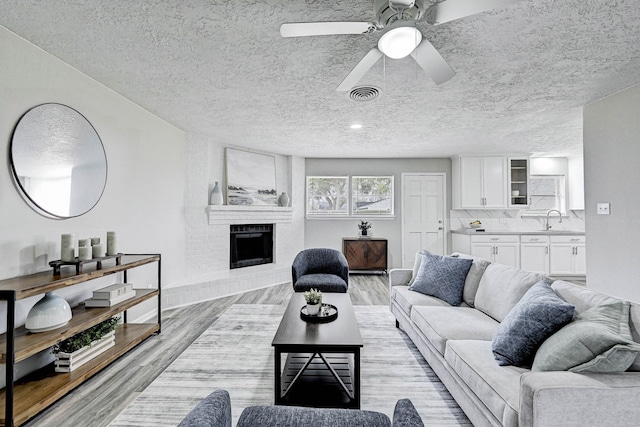 living room with a textured ceiling, a fireplace, sink, light wood-type flooring, and ceiling fan