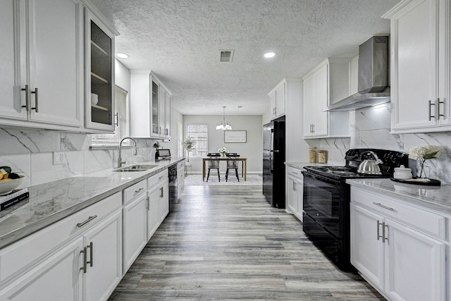 kitchen with decorative light fixtures, wall chimney range hood, black appliances, sink, and white cabinets