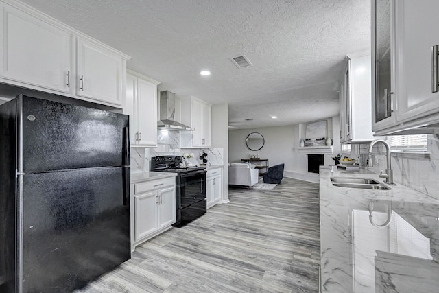 kitchen with light stone countertops, white cabinets, black appliances, wall chimney range hood, and sink