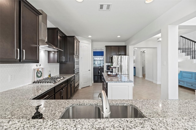 kitchen with stainless steel appliances, tasteful backsplash, wall chimney range hood, light stone countertops, and sink