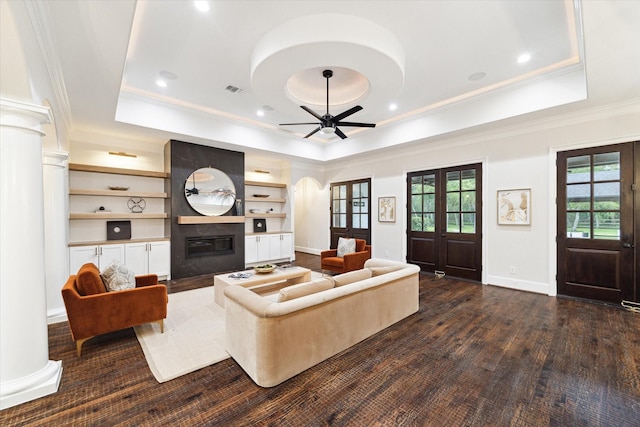 living room with ceiling fan, a large fireplace, a tray ceiling, built in shelves, and dark hardwood / wood-style flooring