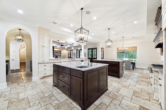 kitchen featuring an island with sink, hanging light fixtures, light stone countertops, dark brown cabinetry, and sink