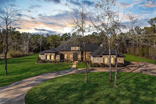 french provincial home with a yard, a forest view, stone siding, and driveway