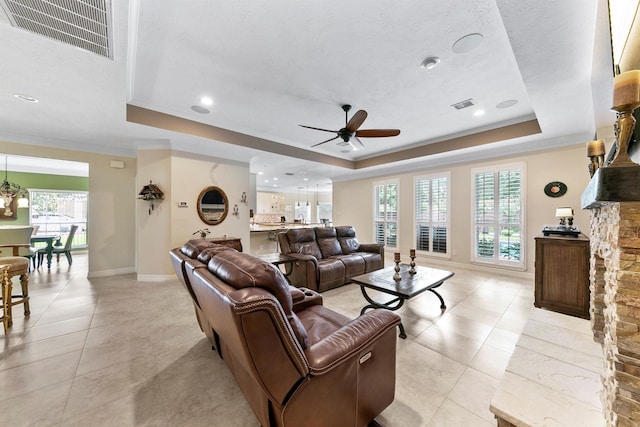 living room with a tray ceiling, a stone fireplace, ornamental molding, and ceiling fan