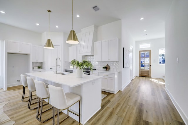 kitchen with white cabinetry, sink, hanging light fixtures, a kitchen island with sink, and custom range hood