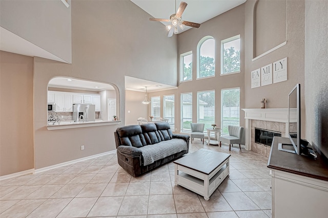 living room featuring ceiling fan, a towering ceiling, a tiled fireplace, and light tile patterned flooring